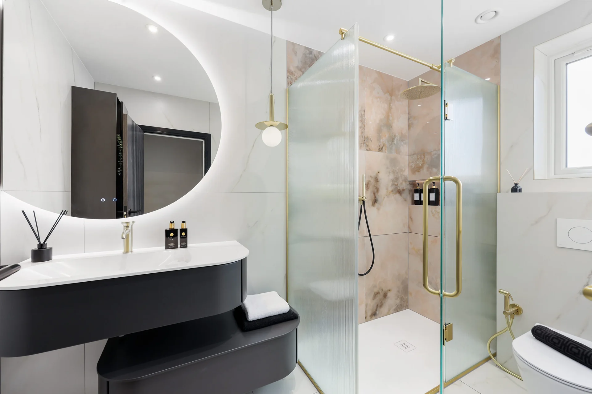 Completed shower room with a black floating vanity, curved backlit mirror, fluted-glass enclosure and warm-metal detailing.