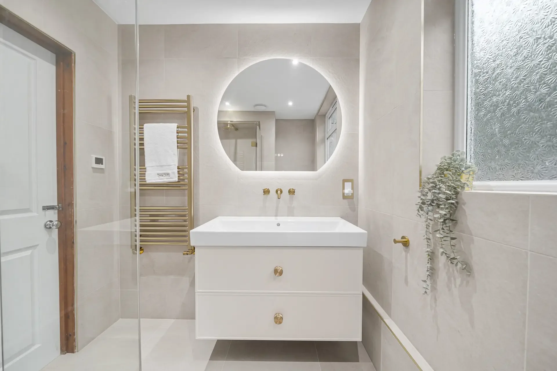 Completed warm-neutral shower room with a floating vanity, rounded backlit mirror and brushed warm-metal details.