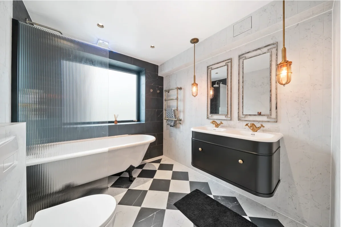 Boutique hotel-style bathroom in Ilford with a freestanding bath, fluted-glass screen, black double vanity and chequerboard floor.